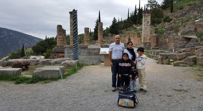 Delphi, Greece A family poses in front of ancient ruins in Delphi. Trees and mountains are visible in the background.